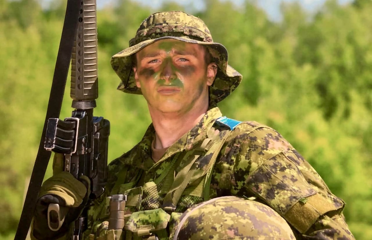 A soldier poses for a photo in military fatigues while holding a gun and his helmet.