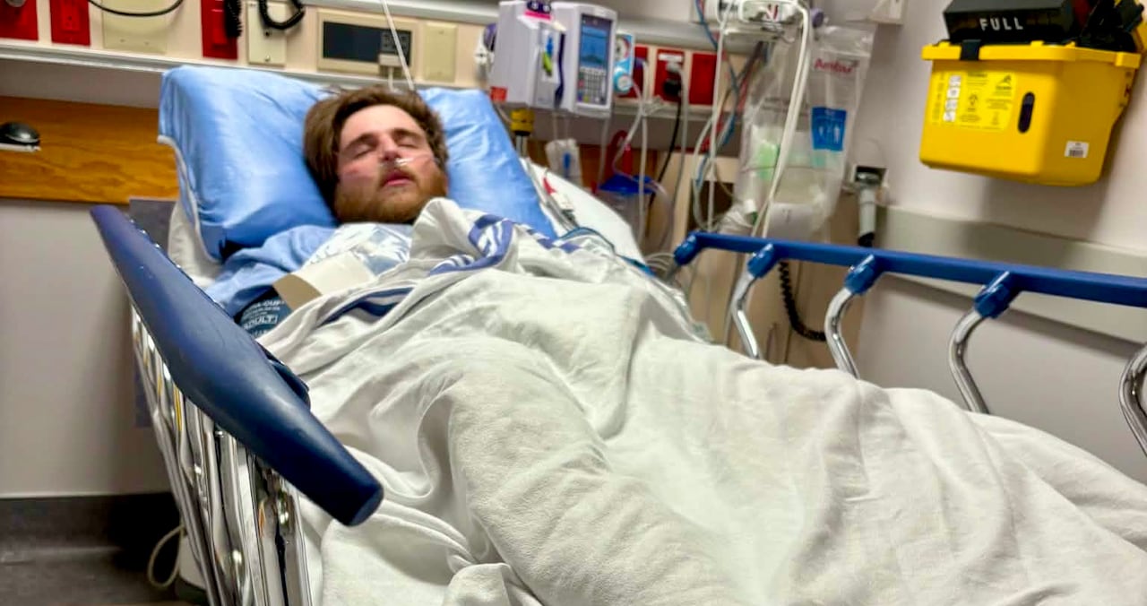 A man lays in a hospital bed covered in a blanket with his eyes closed.