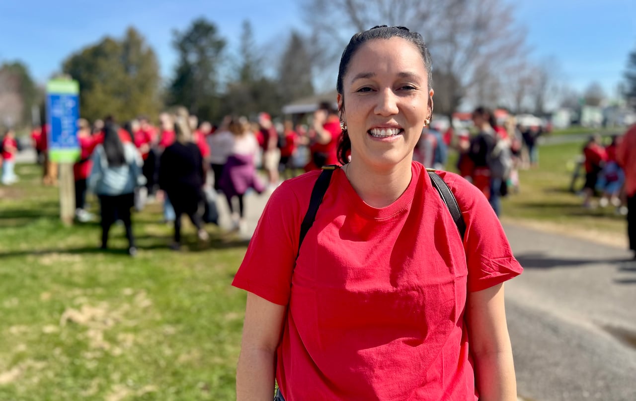 A woman with her hair pulled back, wearing a red t-shirt, smiles at the camera while standing outside on a sunny day.