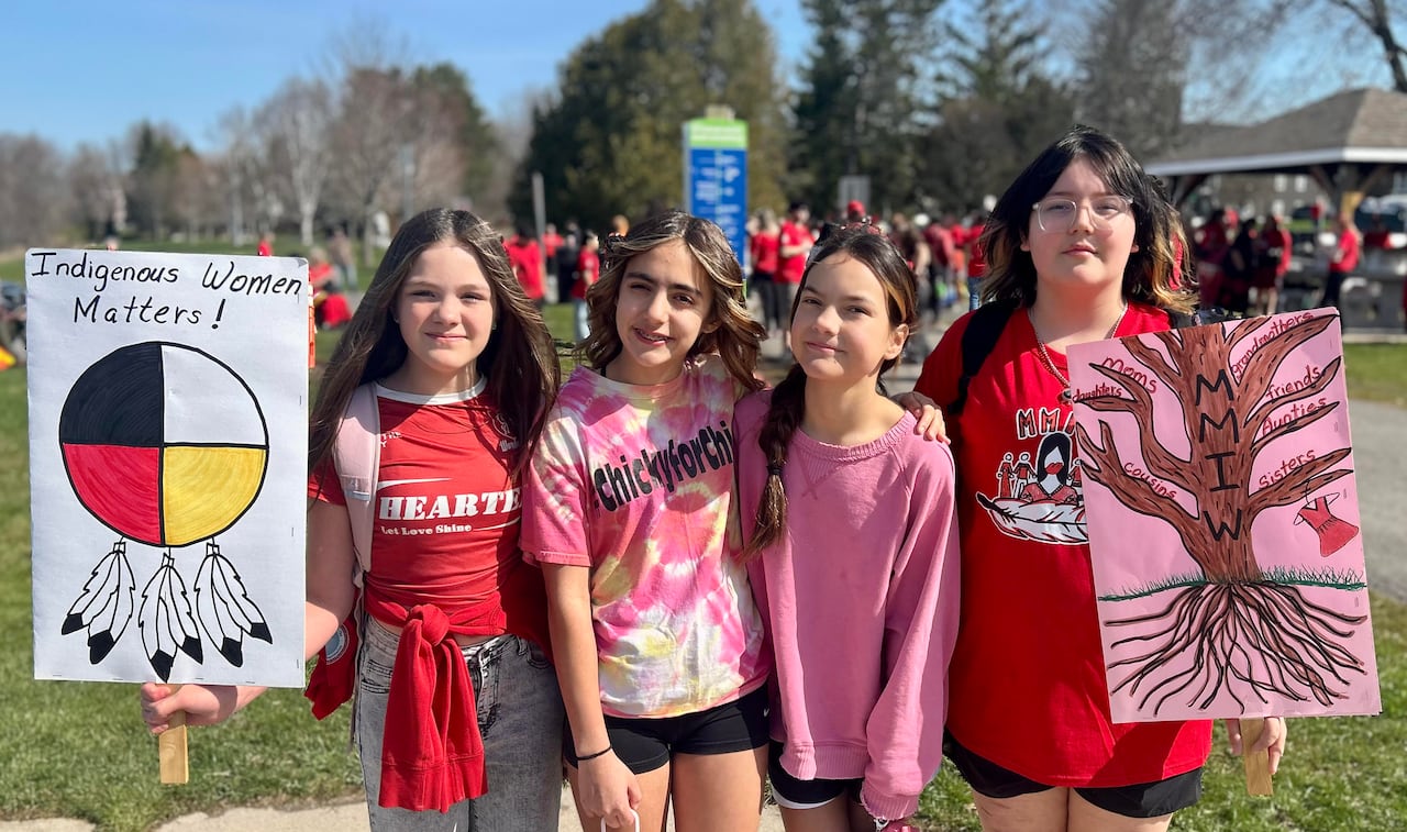 Four young teenagers pose for a photo while holding signs and wearing red and pink, on a sunny day.
