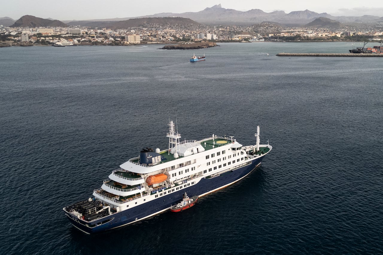An aerial shot of small boat beside a white and blue cruise ship anchored on the blue water offshore, with buildings lining on a mountainous island in the background.