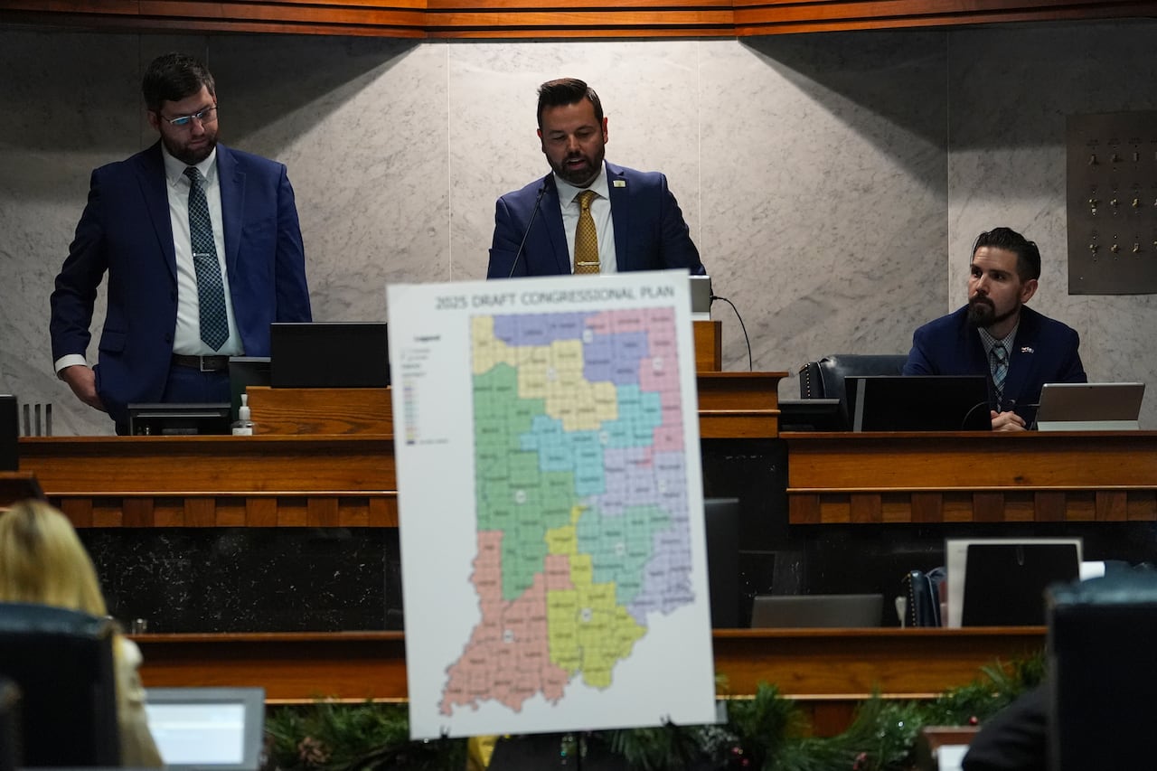A bearded man in a suit and tie standing behind a podium in what appears to be a meeting room or chamber, holds a board that displays a map.