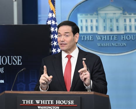 A man in a suit at a podium, speaking and holding up both index fingers.