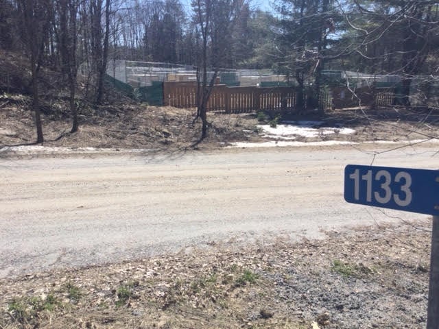 A sign reading 1133 in the foreground features a gate, chain link fence and tarps covering structures across a gravel road  in the distance on the other side.