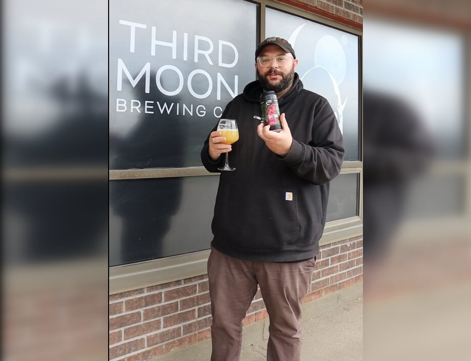A bearded man wearing a hat and glasses is shown with a can of beer and a glass outside of the brewery he co-owns.