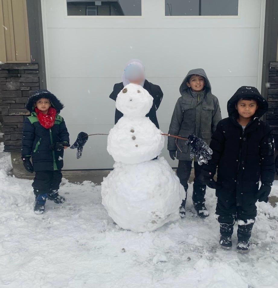 Four young children stand next to and behind a snowman. The girl's face has been blurred.