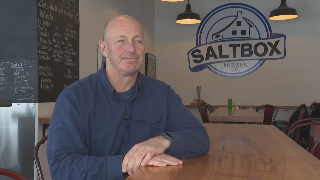 A man sits at a wooden table. Behind him is a sign for Saltbox Brewing Co.
