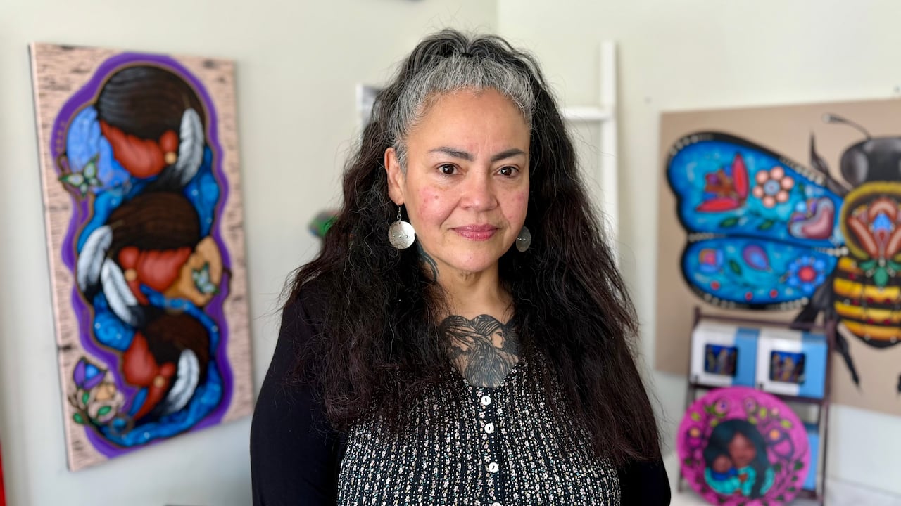 A woman stands in a room with brightly coloured paintings hanging on the wall behind her.