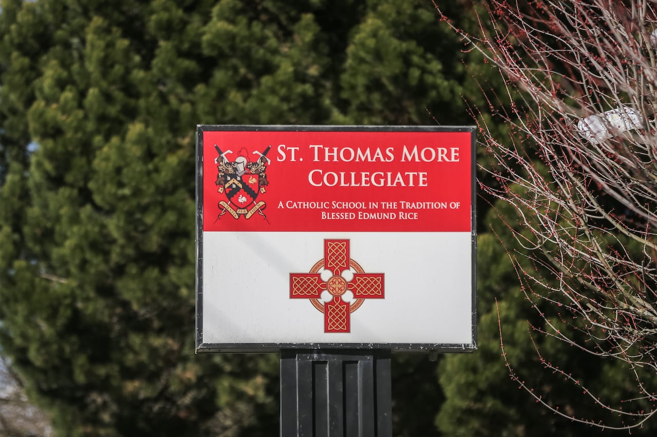 A red and white sign outside St. Thomas More Collegiate, a Catholic school, is pictured in front of evergreen trees.