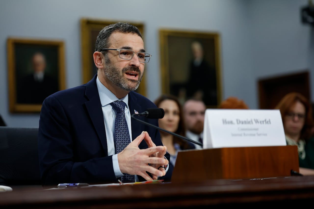 A bearded man wearing a dark suit and tie speaks while seated in what appears to be a courtroom or government chamber.
