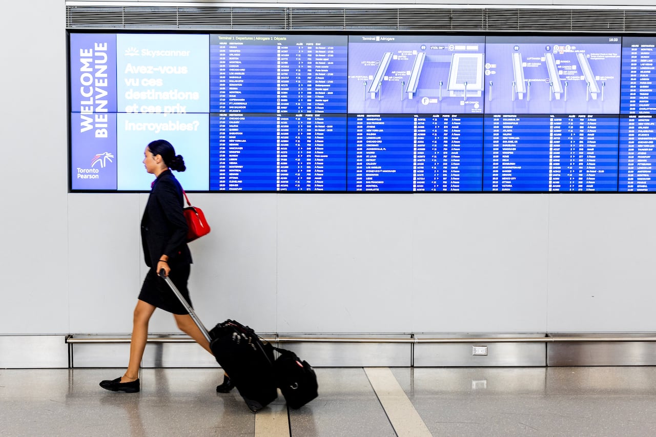Woman in skirt suit pulls luggage in front of electronic board listing flight information.