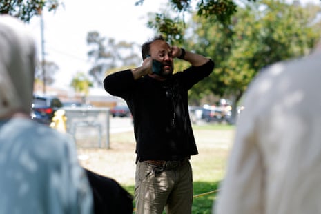 A man in a black shirt speaks on a mobile phone outdoors in a park with blurred figures nearby.