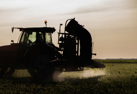 A silhouetted tractor with spray equipment releases a mist over a green field at dusk