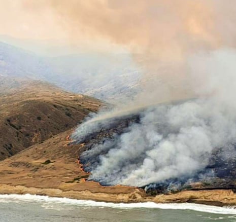 Smoke billows from a hillside fire near a coastline, with flames visible along the burn line